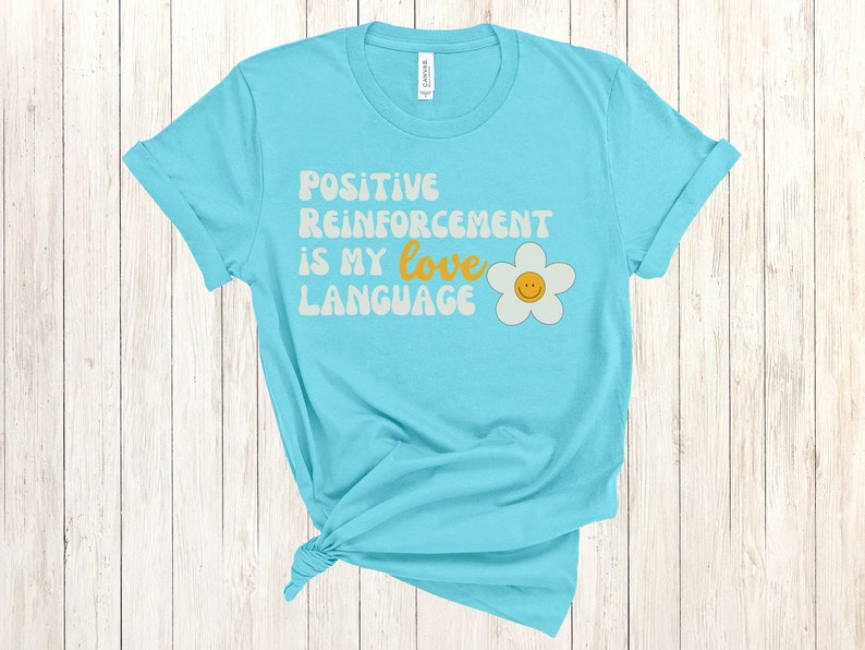 Puede incluir: Una camiseta azul turquesa con el texto "Positive Reinforcement is my love language" en una fuente retro con una margarita blanca con una carita sonriente amarilla.