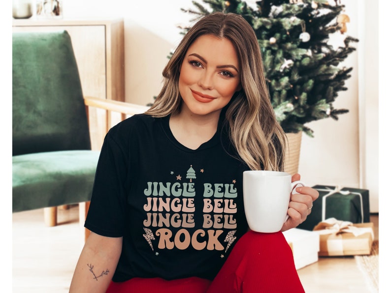 May include: A black t-shirt with a festive Christmas design featuring the text "Jingle Bell Jingle Bell Jingle Bell Rock" in a retro style with a green and pink color scheme.