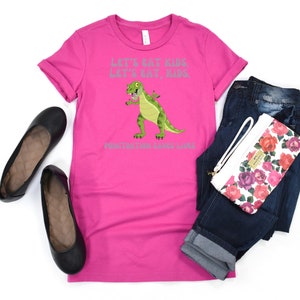 May include: Pink t-shirt featuring a green dinosaur graphic and the text "Let's eat kids, let's eat kids, punctuation saves lives".