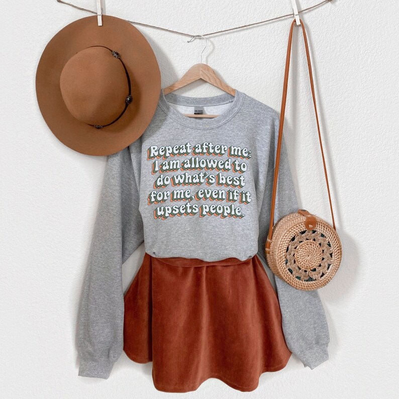 May include: Grey sweatshirt with the text "Repeat after me: I am allowed to do what's best for me, even if it upsets people." The sweatshirt is paired with a brown corduroy skirt and a brown woven bag.