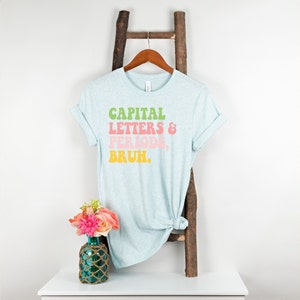 Puede incluir: Una camiseta azul claro con el texto "CAPITAL LETTERS & PERIODS, BRUH." impreso en letras retro verdes, rosas, amarillas y naranjas.