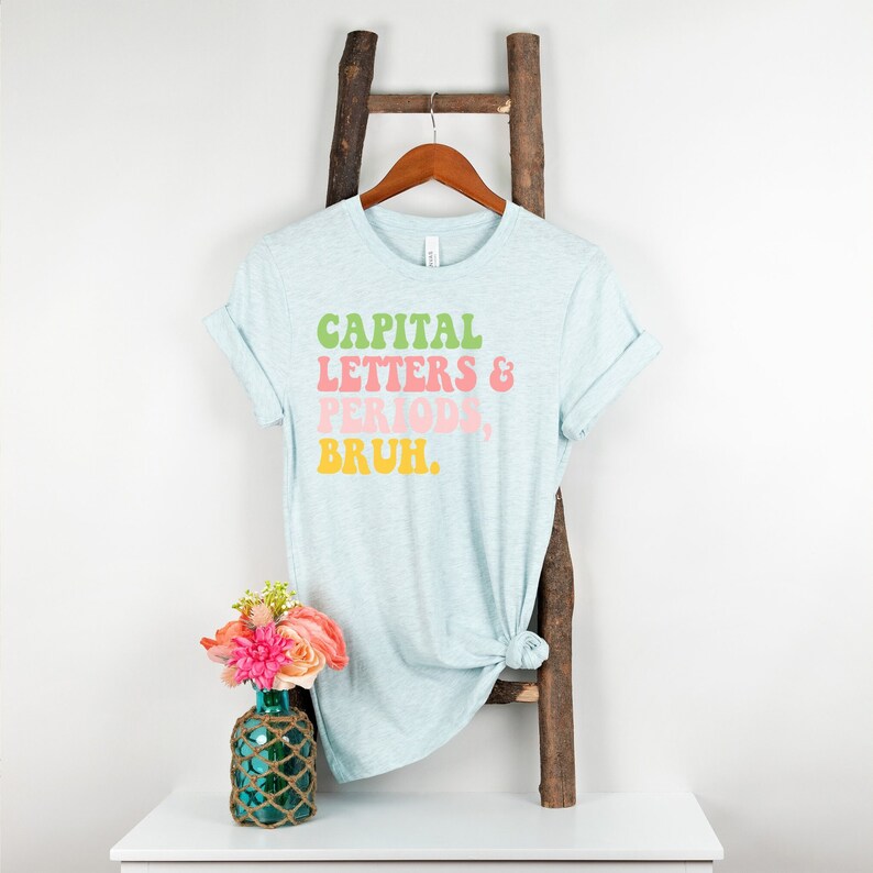 Puede incluir: Una camiseta azul claro con el texto "CAPITAL LETTERS & PERIODS, BRUH." impreso en letras retro verdes, rosas, amarillas y naranjas.