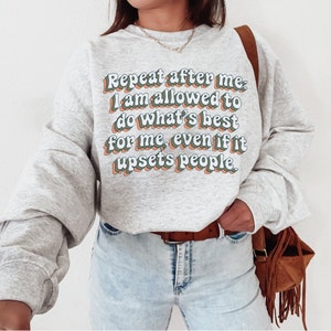 May include: A grey sweatshirt with the text "Repeat after me: I am allowed to do what's best for me, even if it upsets people." printed in a retro font with green, blue, and orange accents.