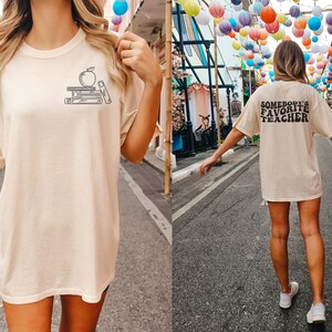May include: Two photos of a person wearing a light beige t-shirt with a graphic of a stack of books and an apple on the front. The second photo shows the back of the shirt with the text "Somebody's Favorite Teacher" printed in black.