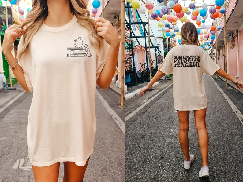 May include: Two photos of a person wearing a light beige t-shirt with a graphic of a stack of books and an apple on the front. The second photo shows the back of the shirt with the text "Somebody's Favorite Teacher" printed in black.
