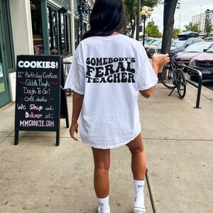 Peut inclure: T-shirt blanc avec un texte noir qui dit "Somebody's Feral Teacher" dans une police de caract&egrave;res r&eacute;tro. La personne portant le t-shirt marche dans une rue de la ville.