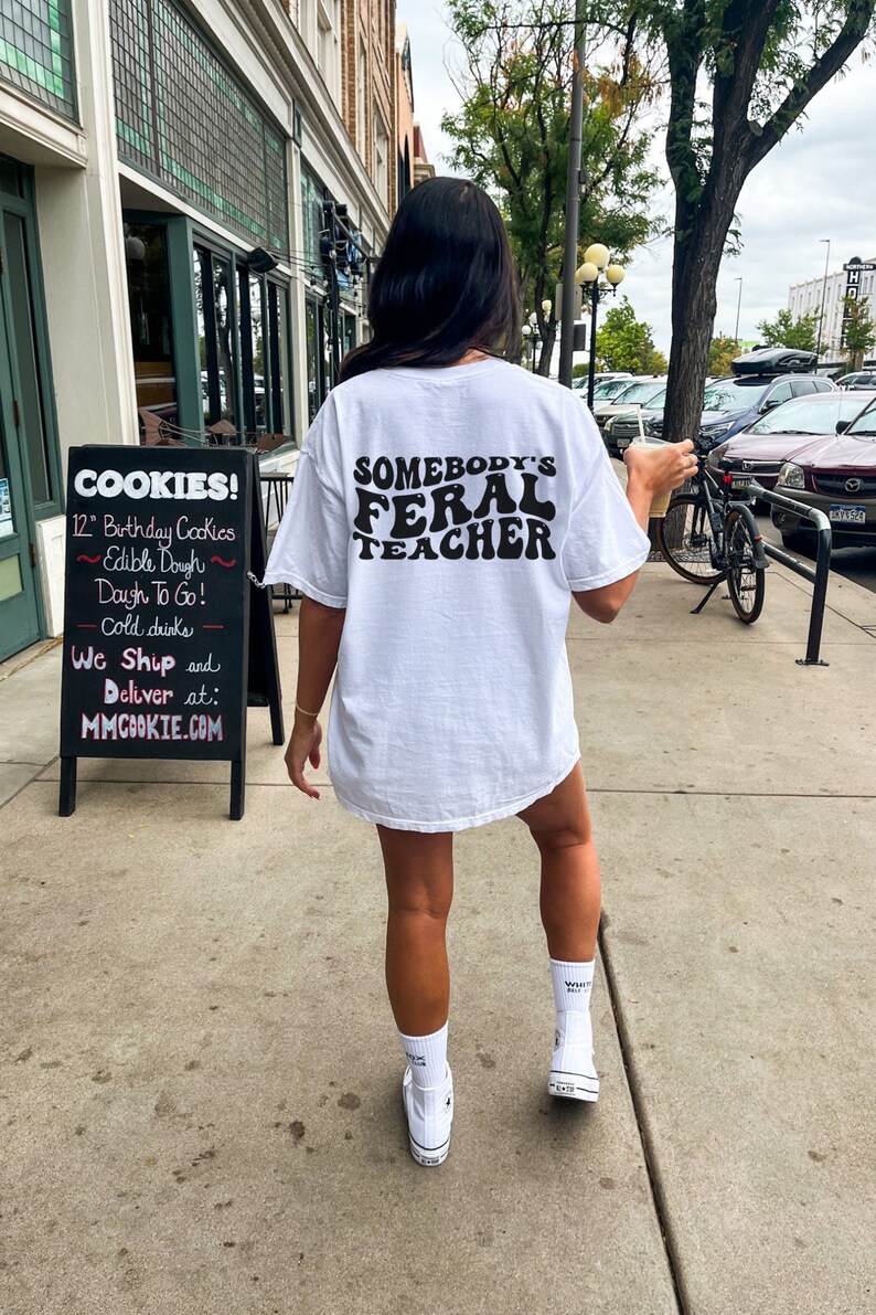 Peut inclure: T-shirt blanc avec un texte noir qui dit "Somebody's Feral Teacher" dans une police de caract&egrave;res r&eacute;tro. La personne portant le t-shirt marche dans une rue de la ville.