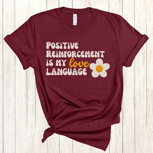 Puede incluir: Camiseta color burdeos con el texto "Positive Reinforcement is my love language" en una fuente retro con una margarita blanca con una cara sonriente amarilla.