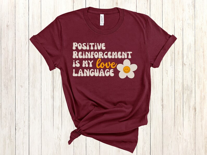 Puede incluir: Camiseta color burdeos con el texto "Positive Reinforcement is my love language" en una fuente retro con una margarita blanca con una cara sonriente amarilla.