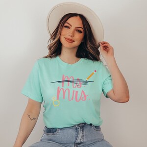 May include: A woman wearing a light blue t-shirt with the text "Mrs." crossed out with a black line and a yellow pencil with a pink eraser above the text. The shirt also has a yellow ring with a pink gem on it. She is wearing blue jeans and a white hat.