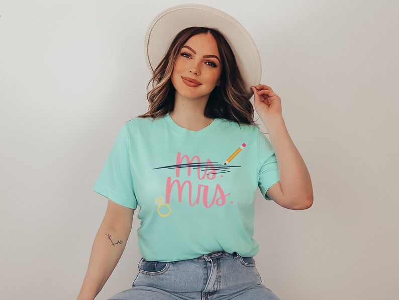May include: A woman wearing a light blue t-shirt with the text "Mrs." crossed out with a black line and a yellow pencil with a pink eraser above the text. The shirt also has a yellow ring with a pink gem on it. She is wearing blue jeans and a white hat.