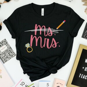 May include: Black t-shirt with the text "Ms. Mrs." in pink script with a diamond ring and a pencil drawn through the "Ms."