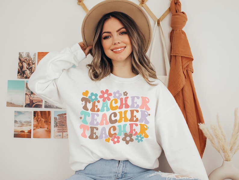May include: White sweatshirt with a colorful retro-style graphic that says "Teacher Teacher Teacher" with flowers and hearts.