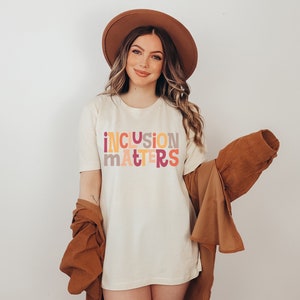 Puede incluir: Una mujer con una camiseta blanca con el texto "inclusion matters" en letras de colores. Lleva un sombrero marr&oacute;n y una chaqueta marr&oacute;n.