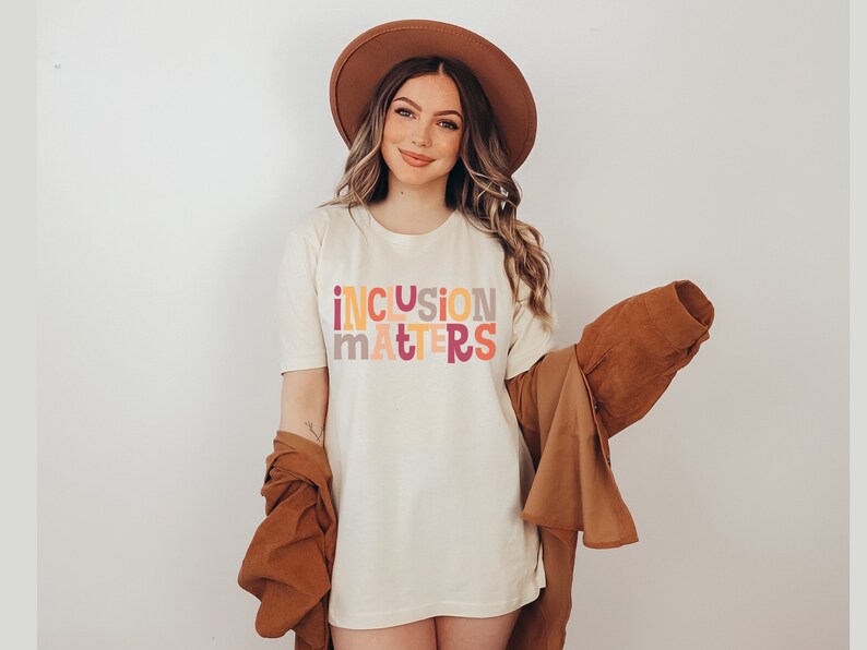 Puede incluir: Una mujer con una camiseta blanca con el texto "inclusion matters" en letras de colores. Lleva un sombrero marr&oacute;n y una chaqueta marr&oacute;n.