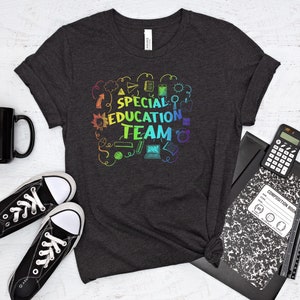 May include: A dark heather grey t-shirt with a rainbow design that says "Special Education Team". The design includes various school-related icons like a pencil, a ruler, a book, and a laptop.