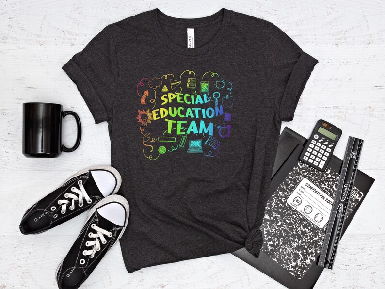 May include: A dark heather grey t-shirt with a rainbow design that says "Special Education Team". The design includes various school-related icons like a pencil, a ruler, a book, and a laptop.