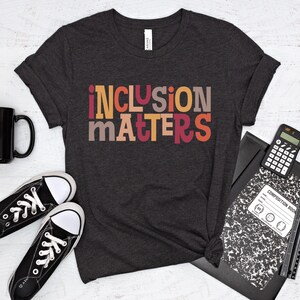 Puede incluir: Una camiseta gris oscuro con el texto "inclusion matters" en letras de bloque de colores. Las letras son en tonos de naranja, rojo, amarillo y marr&oacute;n.