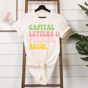 Puede incluir: Una camiseta de color crema con el texto "CAPITAL LETTERS & PERIODS, BRUH." en letras retro de colores.