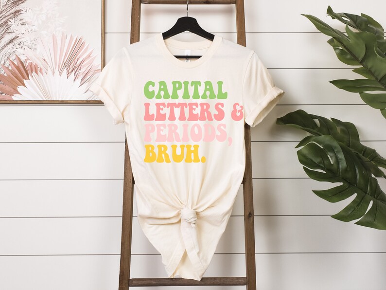 Puede incluir: Una camiseta de color crema con el texto "CAPITAL LETTERS & PERIODS, BRUH." en letras retro de colores.
