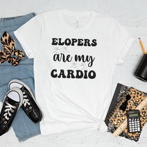 May include: White t-shirt with black text that reads "ELOPERS are my CARDIO". The shirt is laid flat on a white wooden surface with a pair of black sneakers, blue jeans, a leopard print headband, a black mug, a leopard print notebook, a ruler, and a calculator.