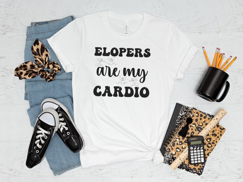 May include: White t-shirt with black text that reads "ELOPERS are my CARDIO". The shirt is laid flat on a white wooden surface with a pair of black sneakers, blue jeans, a leopard print headband, a black mug, a leopard print notebook, a ruler, and a calculator.