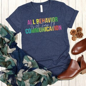 Op de afbeelding: Een marineblauw t-shirt met de tekst "ALL BEHAVIOR IS A FORM OF COMMUNICATION" in regenboogkleuren.