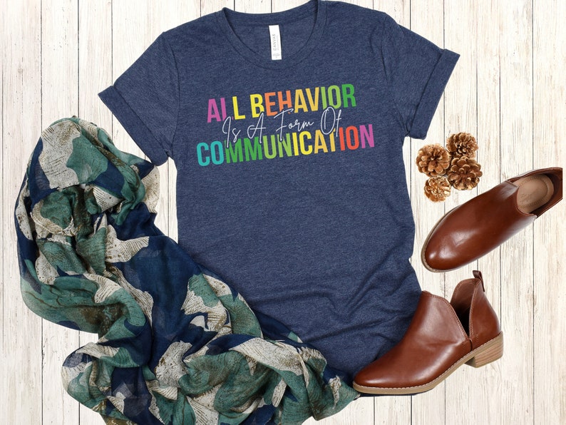 Op de afbeelding: Een marineblauw t-shirt met de tekst "ALL BEHAVIOR IS A FORM OF COMMUNICATION" in regenboogkleuren.