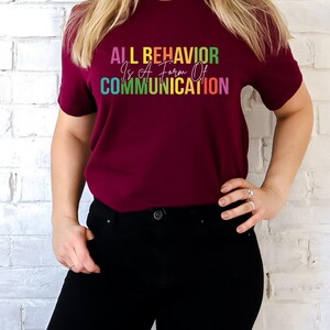 Op de afbeelding: Een maroon t-shirt met de tekst "ALL BEHAVIOR Is A Form Of COMMUNICATION" in regenboogkleuren.