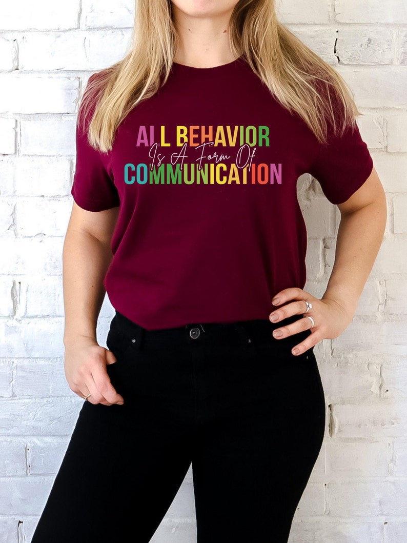 Op de afbeelding: Een maroon t-shirt met de tekst "ALL BEHAVIOR Is A Form Of COMMUNICATION" in regenboogkleuren.