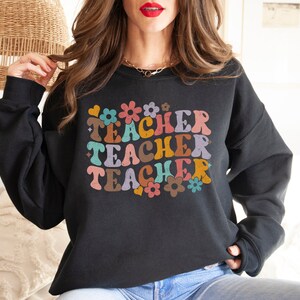 May include: Black sweatshirt with a retro-style graphic that says "Teacher" three times in colorful letters and flowers.