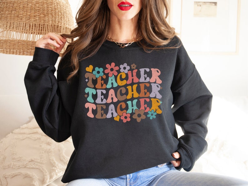 May include: Black sweatshirt with a retro-style graphic that says "Teacher" three times in colorful letters and flowers.