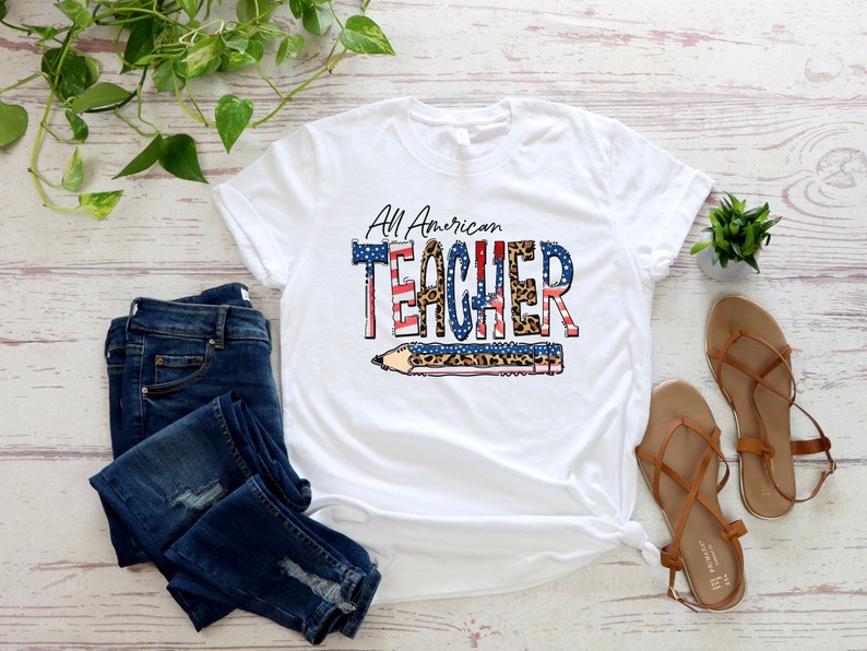 May include: White t-shirt with the text "All American Teacher" in red, white, and blue with a leopard print design. The text is over a pencil graphic with an American flag design.