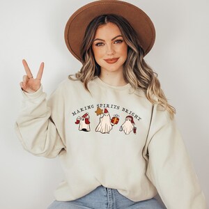 May include: A beige sweatshirt featuring a Christmas-themed design of three ghosts wearing Santa hats. The text "Making Spirits Bright" is printed above the ghosts.