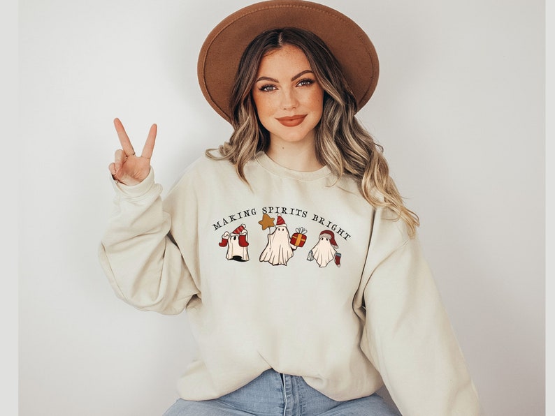 May include: A beige sweatshirt featuring a Christmas-themed design of three ghosts wearing Santa hats. The text "Making Spirits Bright" is printed above the ghosts.