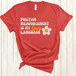 Puede incluir: Camiseta roja con el texto "POSITIVE REINFORCEMENT IS MY love LANGUAGE" en blanco con una margarita blanca con una carita sonriente amarilla en el centro.