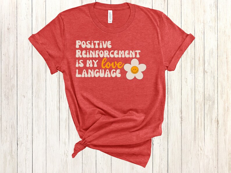 Puede incluir: Camiseta roja con el texto "POSITIVE REINFORCEMENT IS MY love LANGUAGE" en blanco con una margarita blanca con una carita sonriente amarilla en el centro.