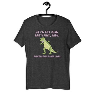 May include: A dark heather grey t-shirt featuring a cartoon dinosaur and the text "Let's eat kids. Let's eat, kids. Punctuation saves lives."