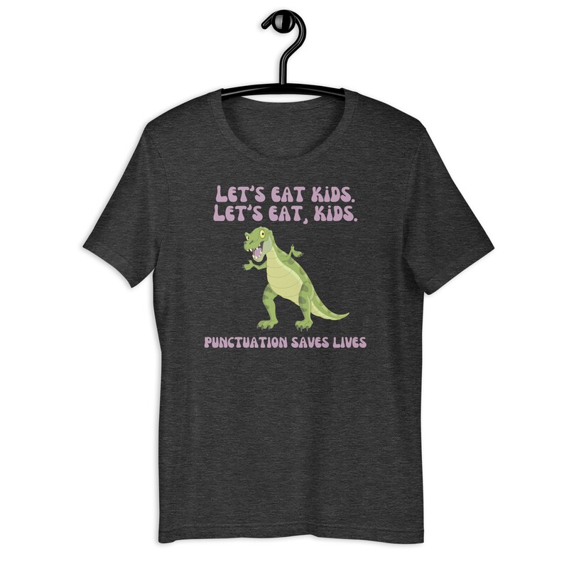 May include: A dark heather grey t-shirt featuring a cartoon dinosaur and the text "Let's eat kids. Let's eat, kids. Punctuation saves lives."