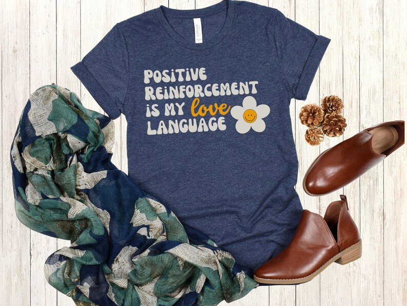Puede incluir: Una camiseta azul marino con el texto "POSITIVE REINFORCEMENT IS MY love LANGUAGE" en una fuente retro con una margarita amarilla con una cara sonriente.