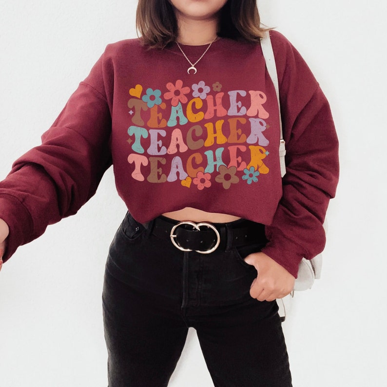 May include: A maroon cropped sweatshirt with the text "TEACHER TEACHER TEACHER" in colorful retro lettering with flowers and hearts. The sweatshirt is worn by a person with black jeans and a silver belt.