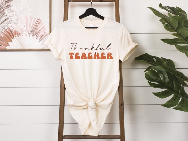 Puede incluir: Una camiseta de color crema con el texto "Thankful Teacher" en letras marrones y naranjas. La camiseta se muestra en una escalera de madera con un fondo blanco.