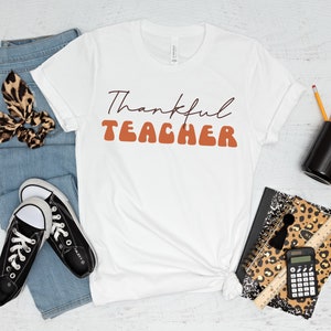 Puede incluir: Camiseta blanca con el texto "Thankful Teacher" en letras marrones y naranjas. La camiseta est&aacute; colocada sobre una superficie de madera blanca con jeans, zapatillas, una libreta con estampado de leopardo, l&aacute;pices y una taza.