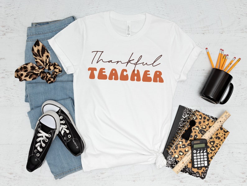Puede incluir: Camiseta blanca con el texto "Thankful Teacher" en letras marrones y naranjas. La camiseta est&aacute; colocada sobre una superficie de madera blanca con jeans, zapatillas, una libreta con estampado de leopardo, l&aacute;pices y una taza.