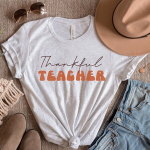 Puede incluir: Una camiseta blanca con el texto "Thankful Teacher" en letras naranja y marr&oacute;n. La camiseta est&aacute; colocada sobre una superficie marr&oacute;n con un sombrero, unos vaqueros y otros accesorios.