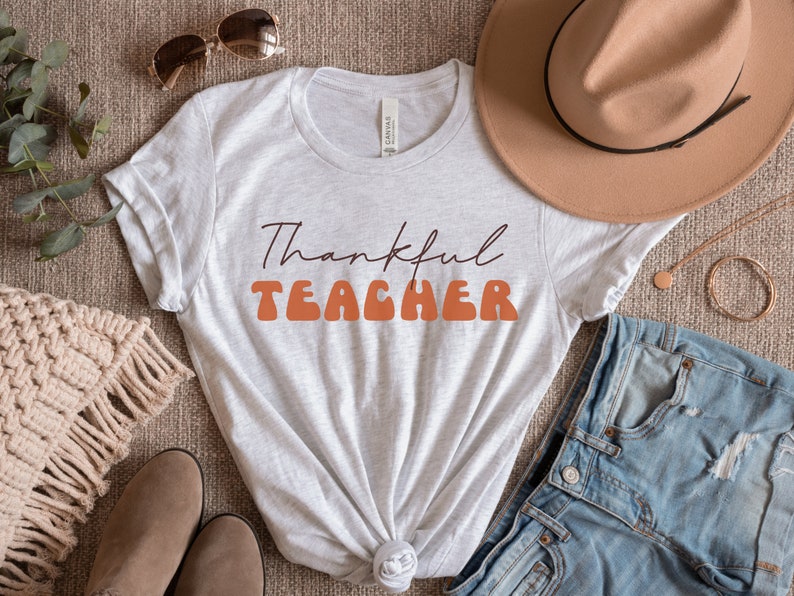 Puede incluir: Una camiseta blanca con el texto "Thankful Teacher" en letras naranja y marr&oacute;n. La camiseta est&aacute; colocada sobre una superficie marr&oacute;n con un sombrero, unos vaqueros y otros accesorios.