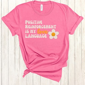 Puede incluir: Camiseta rosa con una margarita blanca y el texto "POSITIVE REINFORCEMENT IS MY LOVE LANGUAGE" en una fuente retro.