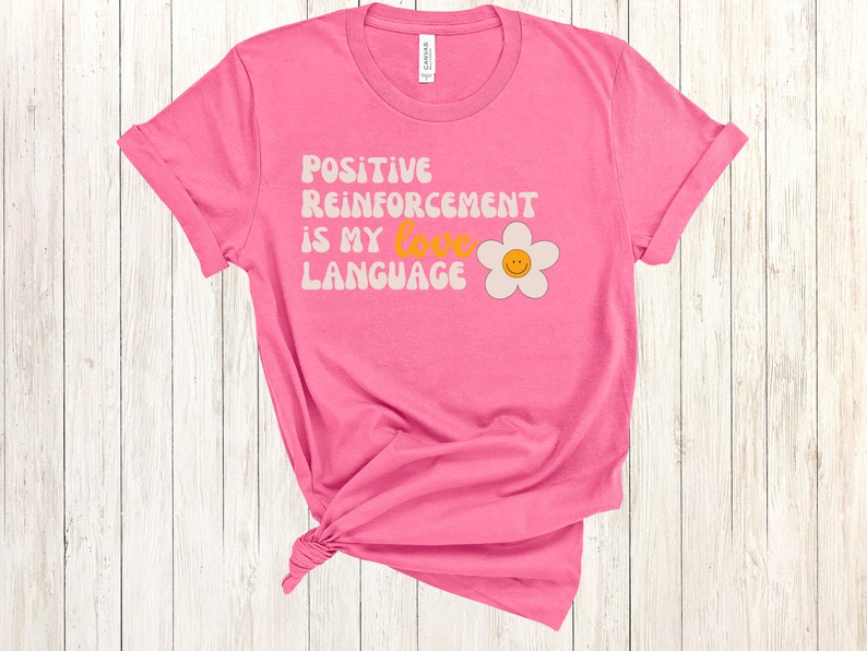 Puede incluir: Camiseta rosa con una margarita blanca y el texto "POSITIVE REINFORCEMENT IS MY LOVE LANGUAGE" en una fuente retro.
