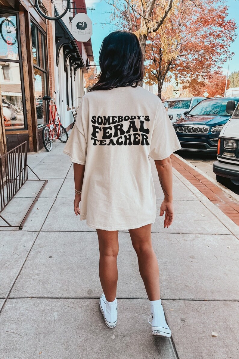 Peut inclure: Un t-shirt blanc surdimensionn&eacute; avec le texte "Somebody's Feral Teacher" imprim&eacute; en noir au dos. Le t-shirt est port&eacute; par une personne qui marche sur un trottoir.
