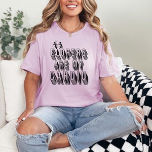May include: A woman wearing a light purple t-shirt with the text "ELOPERS ARE MY CARDIO" in black. The text is in a 3D style with a black outline. She is sitting on a white couch with a black and white checkered throw blanket.